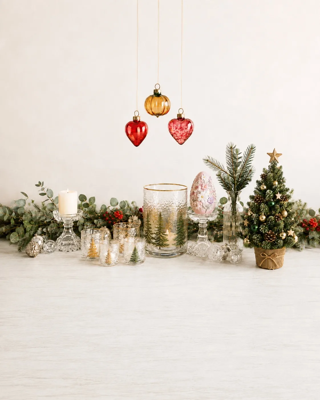 Glass baubles, tree-etched votives, a gold-rimmed hurricane, a footed candle stand and a miniature decorated tree arranged on a pale table with eucalyptus and pine